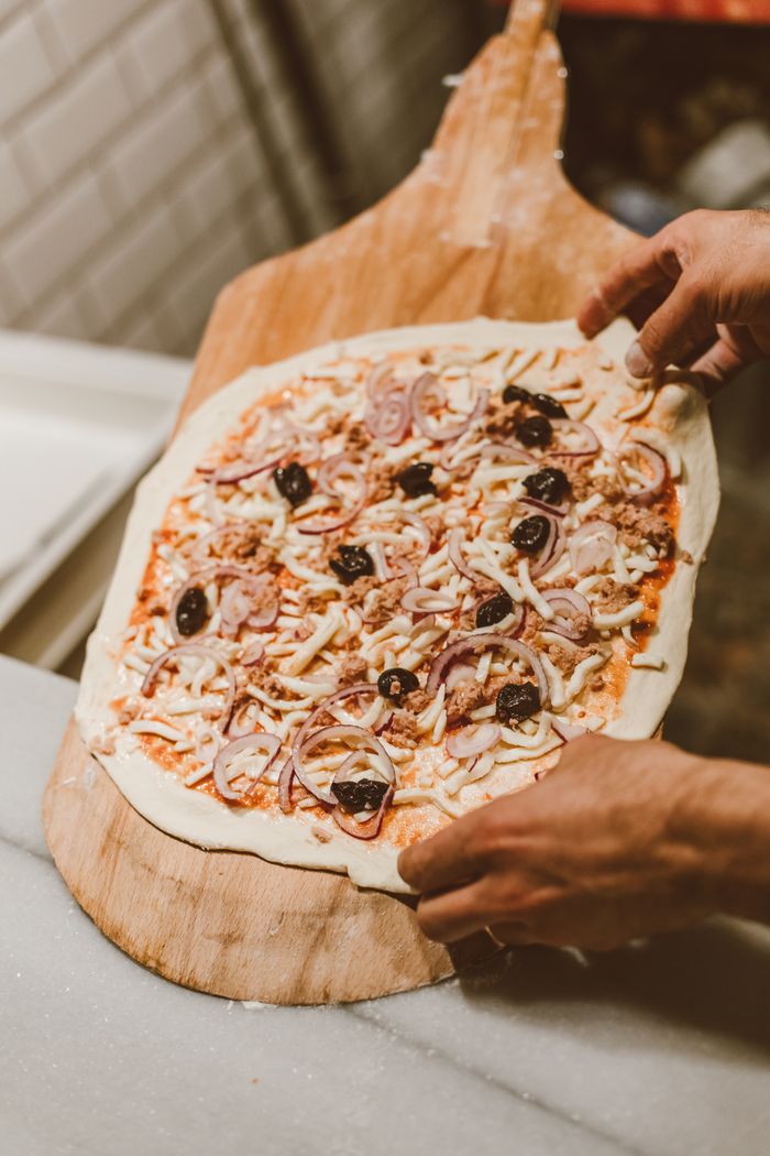 Hands placing a freshly topped pizza on a wooden paddle, ready for baking.